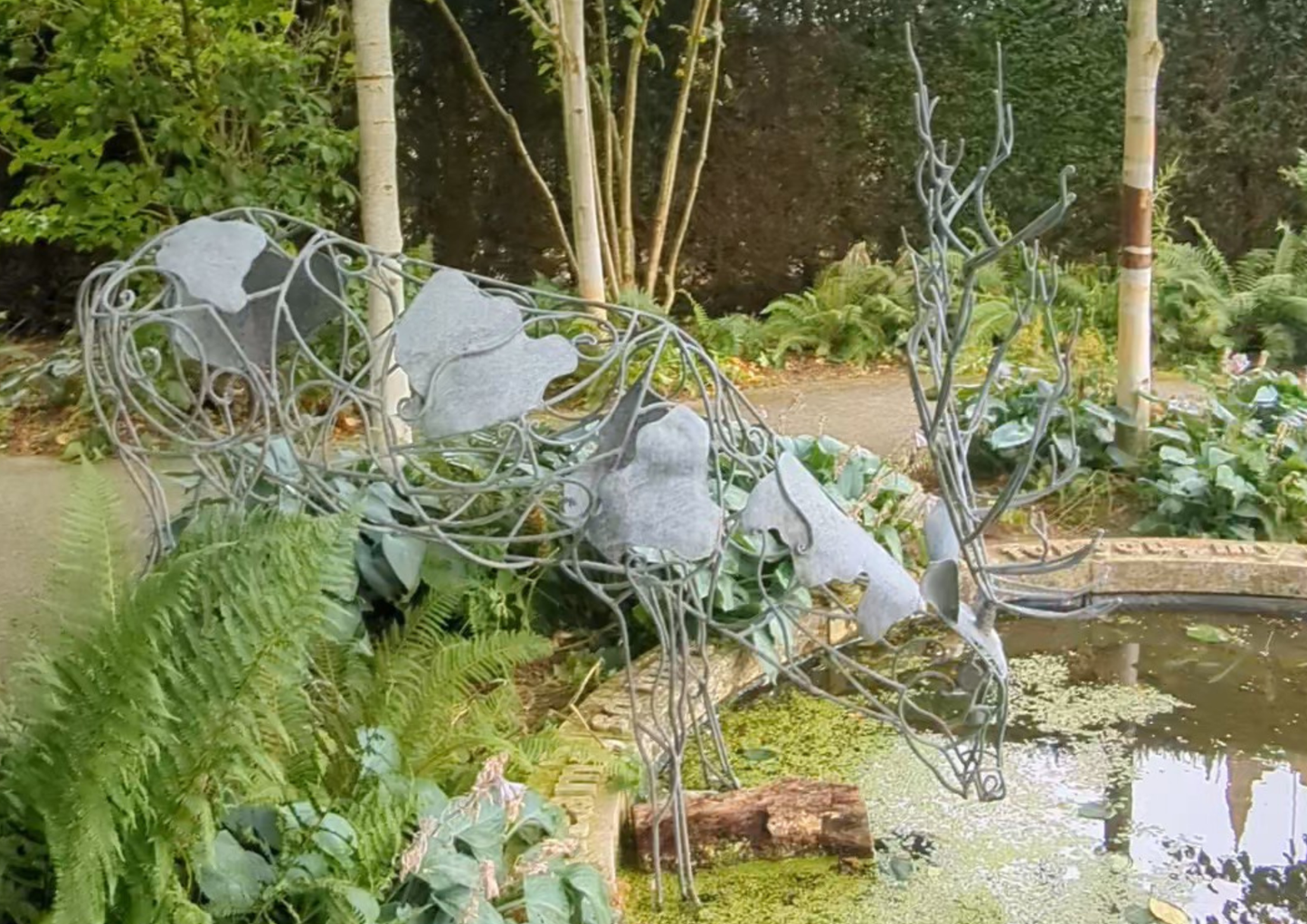 Metallic deer sculpture in a garden setting with plants and a pond.