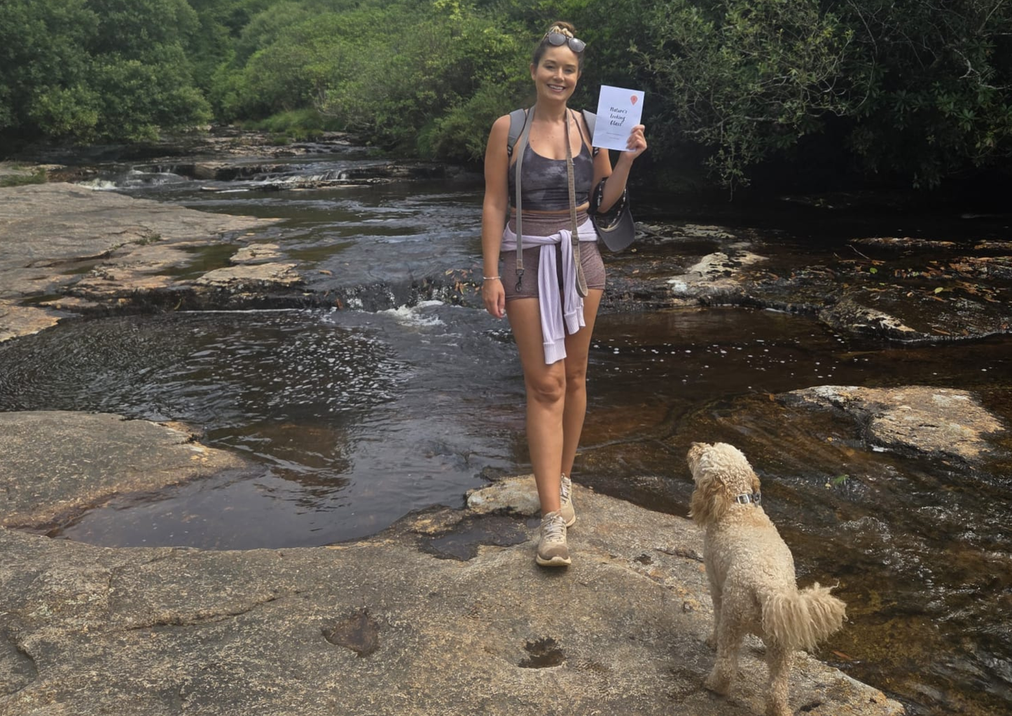 Woman standing on a rocky stream bank with a dog, holding a Snapshot Trips booklet