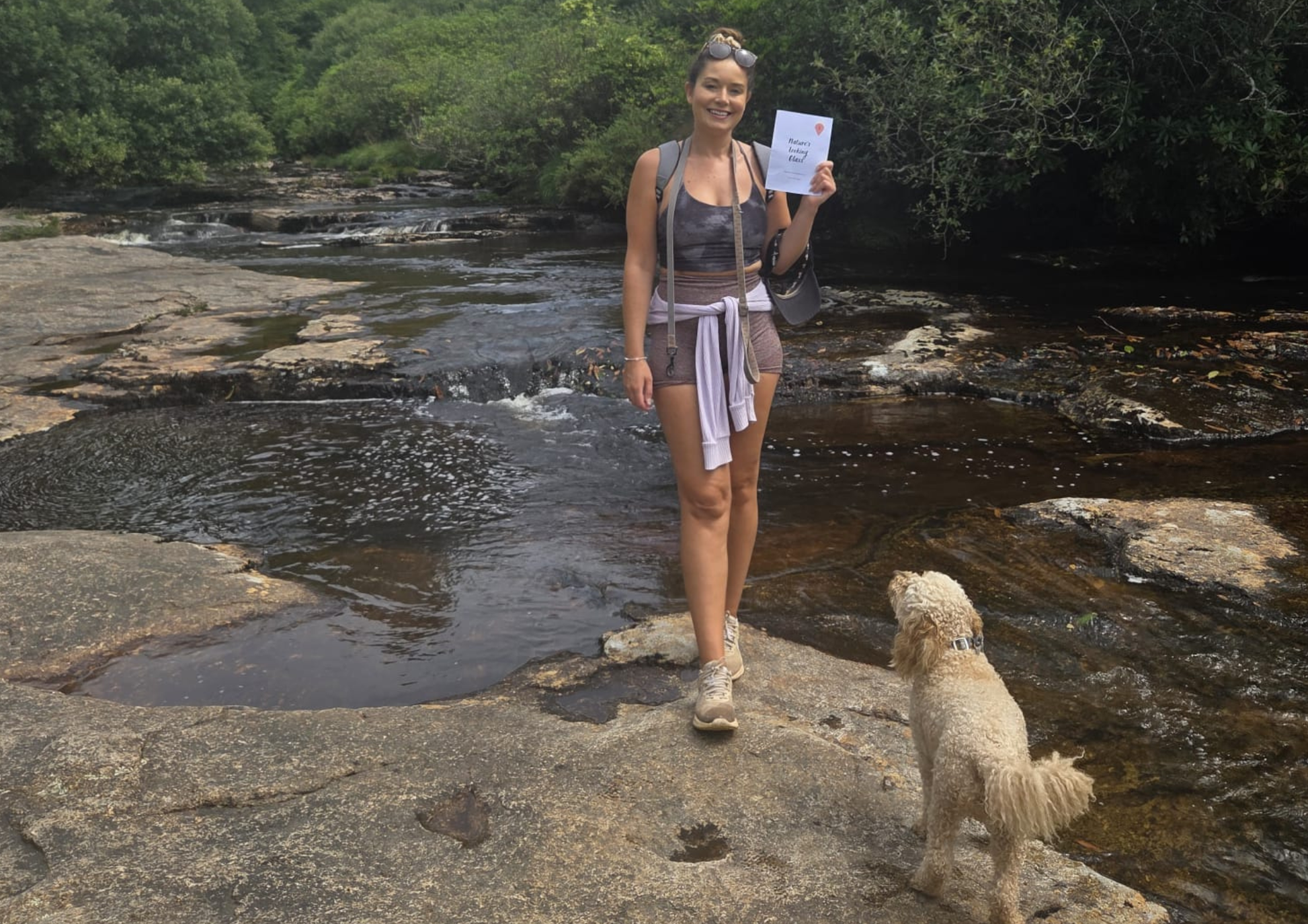 Woman standing on a rocky stream bank with a dog, holding a Snapshot Trips booklet