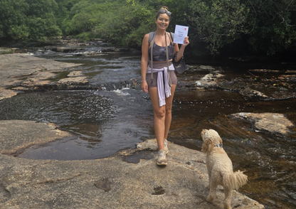 Woman standing on a rocky stream bank with a dog, holding a Snapshot Trips booklet